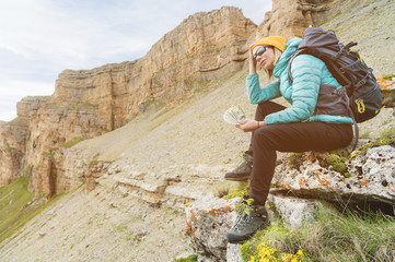 A traveler girl wearing a hat and sunglasses holds a hundred dollar bills in the hands of a fan against the backdrop of cliffs on nature. Keep your head from high cost