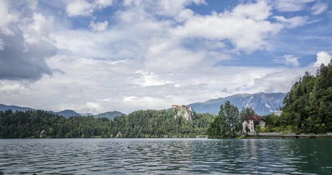 Time lapse of lake Bled with ducks and swans in the water, traffic on the right side, castle on the cliff in the center, and lovely white clouds above with alps in the background.