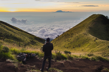 A drone pilot is flying quadrocopter on the mountain above the clouds. Sunrise at mount Merbabu, Indonesia, above the clouds. Volcano Lawu is at the background. Shooting aerial footage.
