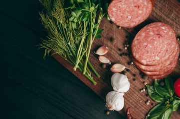 Smoked salami on a black table and a brown board. Sausages with rosemary, garlic and pepper and tomato cherry, dill, parsley.