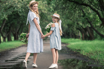 happy stylish mother and daughter holding hands and walking in green park