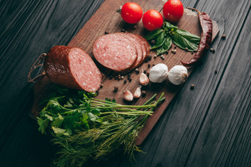 Smoked salami on a black table and a brown board. Sausages with rosemary, garlic and pepper and tomato cherry, dill, parsley.