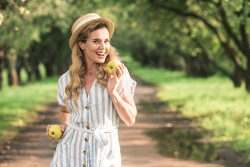 beautiful stylish woman in straw hat holding two apples in orchard