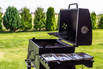 Opened grill with coal in inside, standing on a home garden in the background lawn and thujas.