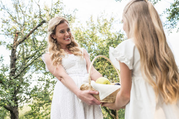 beautiful blonde mother and daughter holding wicker basket with fruits in orchard