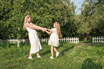 Naklejka premium beautiful happy mother and daughter holding hands and twisting in garden with white fence