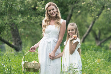 Obraz premium beautiful mother and daughter holding wicker basket with fruits in orchard