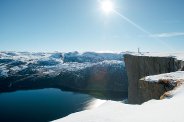 Young traveller woman standing on the Preikestolen cliff edge with raised hands and looking at the mountains. Sunny winter day in Norway. Pulpit Rock