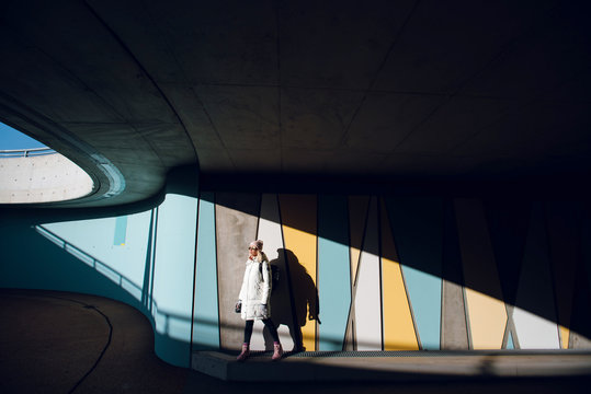 Fototapeta Young traveler woman in white winter coat standing against colorful wall with geometric pattern under the bridge in Norway. Urban background. Art concept.