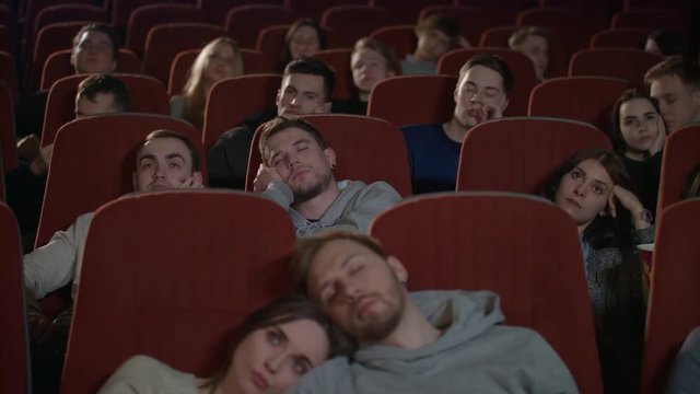 People watch boring movie at cinema in slow motion. Group of young people sleeping watching boring movie in cinema. Bored audience watching film in cinema. Spectators fall asleep from boring film