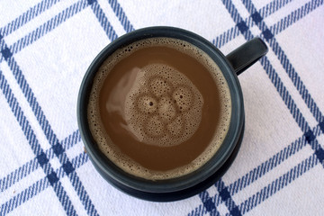 On a white checkered tablecloth there is a cup of coffee. A flower pattern is painted on the surface of coffee.