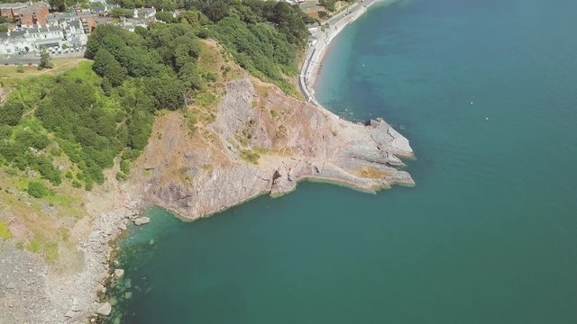 Zooming out with the drone over the landscape of Torquay. The beach and cliffs are visible below. Buildings next to woodland are visible as well.