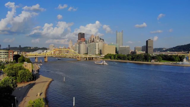 A reverse slowly rising aerial establishing wide shot of the skyline of Pittsburgh, Pennsylvania on an early summer evening. Gateway Clipper riverboat carries tourists in the distance.  	