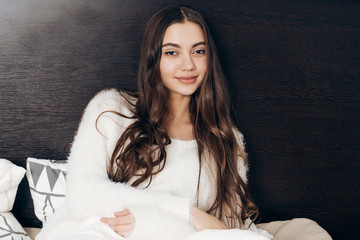 beautiful long-haired girl in white pajamas sits in bed, resting and wants to sleep