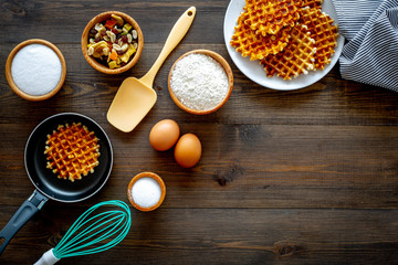 Preparing belgian waffles. Waffle on pan near whisk for whipping and ingrdients. Eggs, flour, sugar, dried fruits on dark wooden background top view copy space