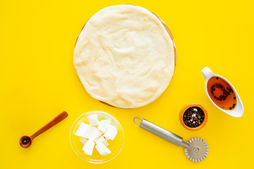 Raw rolled out pizza dough near cheese mozzarella, olive oil, spices, knife for pizza on yellow background top view mockup copy space