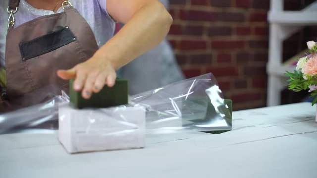 florist girl preparing floral foam for beatiful flower composition in wooden box in design studio. master in apron cutting transparent sheet with scissors. Floristry, handmade, small business concept