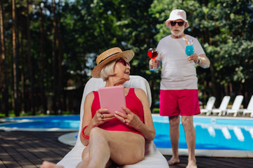 Fototapeta na wymiar Cocktails for wife. Caring retired man bringing some summer cocktails for wife sunbathing near the pool