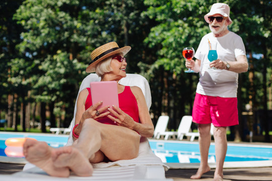 Straw hat. Stylish mature woman wearing straw hat sunbathing near the pool and reading electronic book - Powered by Adobe