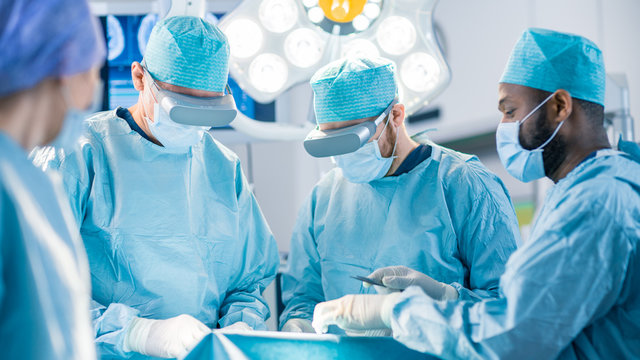 Surgeons Wearing Augmented Reality Glasses Perform State of the Art Mixed Reality Surgery in High Tech Hospital. Doctors and Assistants Working in Operating Room.