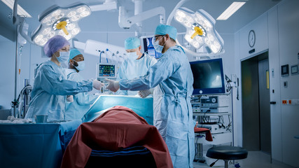 Diverse Team of Professional Surgeons Performing Invasive Surgery on a Patient in the Hospital Operating Room. Nurse Hands Out Instruments to surgeon, Anesthesiologist Monitors Vitals.