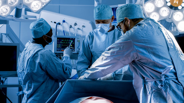 Diverse Team of Professional Surgeons Performing Invasive Surgery on a Patient in the Hospital Operating Room. Nurse Hands Out Instruments to surgeon,  Anesthesiologist Monitors Vitals.