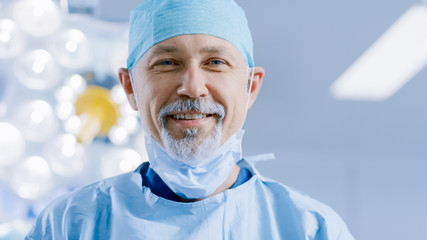 Portrait of the Professional Surgeon Takes off Surgical Mask after Successful Operation Simling Into Camera. In the Background Modern Hospital Operating Room.