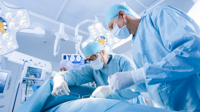 Low Angle Shot of a Diverse Team of Professional Surgeons Performing Invasive Surgery on a Patient in the Hospital Operating Room. Surgeons Use Instruments