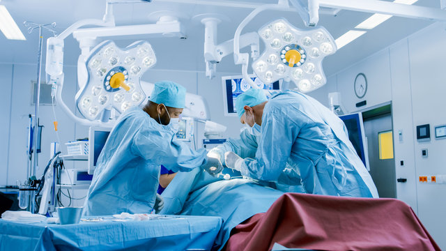 Diverse Team Of Professional Surgeons Performing Invasive Surgery On A Patient In The Hospital Operating Room. Surgeons Use Instruments.