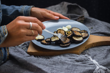 Woman hands sticks on fork roasted eggplants slices fro stylish grey plate. Sprinkled with herbs, black pepper and garlic powder. Perfect vegan dish. No refined oil. Vegetarian lunch. Small portion