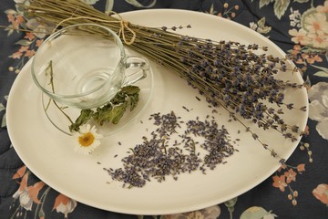 Glass cup and bouquet of dry lavender on a white earthenware dish, floral fabric background