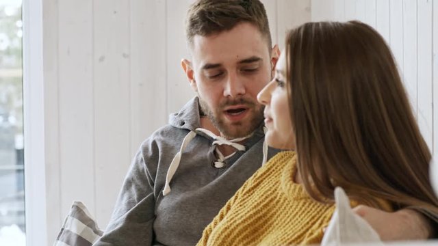 Handheld shot of loving bearded man talking and looking with affection at his laughing girlfriend while relaxing with her on sofa