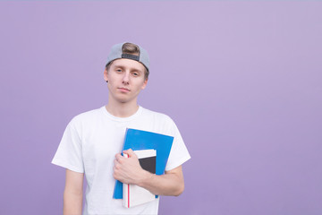Serious young man in a white T-shirt stands on a purple background with books and notebooks in his...