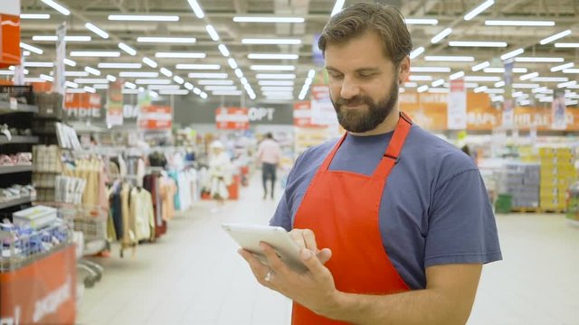Handsome supermarket clerk with beard using a touch screen tablet in supermarket, he is smiling at camera