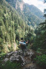  a young guy with a tourist backpack looks down from the mountain to the river and a thick green forest