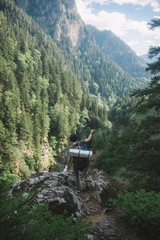  a young guy with a tourist backpack looks down from the mountain to the river and a thick green forest