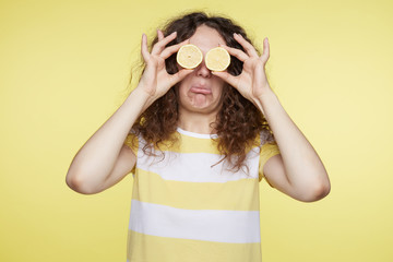 Curly haired funny woman closes eyes with two halves of lemons and opens mouth demonstrating sour taste of fruit, isolated over yellow background. Unhappy young female makes grimace or pulls face.