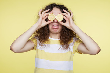 Curly haired funny woman closes eyes with two halves of lemons and opens mouth, shows tongue, demonstrating sour taste of fruit, isolated over yellow background. Negative emotion and feelings concept.