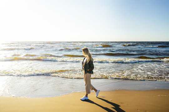 side view of young woman in stylish clothing walking on seashore on summer day