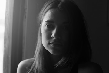 Black and white natural close up portrait of mixed raced woman, looking deeply at camera, posing indoors under window light. Candid shot of young sensual female with no make up and no retouch.