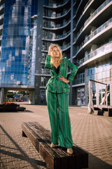 Beautiful girl in a green suit on a blurred building background. Stylish blonde girl. Fashionable girl. Everyday life of a beautiful girl. Lovely blonde in a stylish suit.