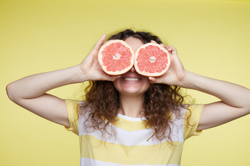 Close up isolated portrait of young curly haired woman holding halved oranges at her eyes. Headshot of funny girl wearing stripe T-shirt poses on yellow wall. Human face expressions and emotions.