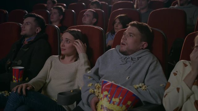 Young guy scared while watching horror film in movie theatre. Young man sprinkling popcorn in slow motion at cinema. Spectators frightened by horror film. People watching scary movie
