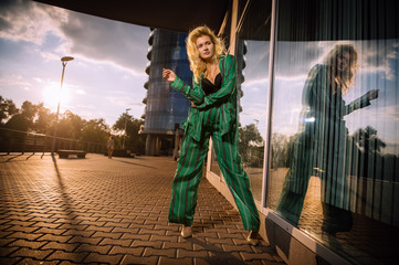 A beautiful girl in a green suit stands near a glass wall at sunset in the city. Stylish blonde. The girl is reflected in the mirror surface. Everyday life of a beautiful girl. Beautiful blonde.
