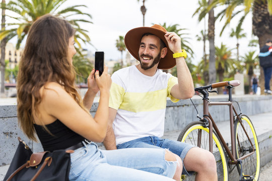 Spain, Barcelona, happy couple sitting on bench taking a smartphone picture
