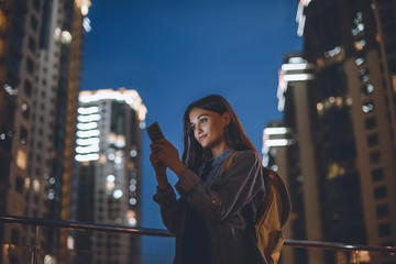 smiling woman with backpack using smartphone on street with night city lights on background