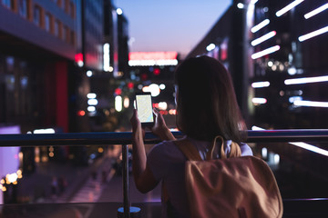 rear view of woman with backpack and smartphone with map on screen in hands standing on night city street