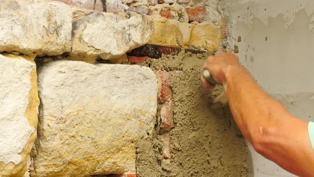 Detail of worker man hands putting fresh mortar on wall. Building stony and bricks wall with mortar in construction site