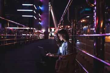 side view of young woman using laptop on street with night city lights on background