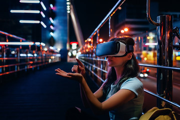Fototapeta premium young woman in virtual reality headset sitting on street with night city on background